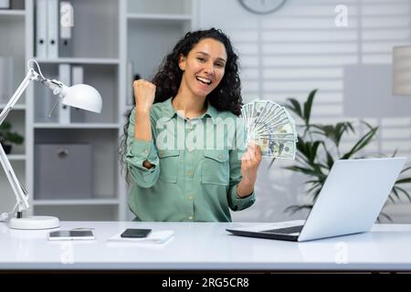 A young Latin American woman is sitting at the desk in the office and holding cash money in her hands. Rejoices and smiles at the camera, shows a victory hand gesture yes. Stock Photo