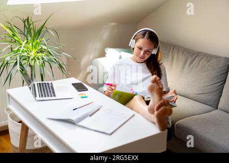 Young woman university student sitting at the table at home with feet up and reading student's textbook. Stock Photo