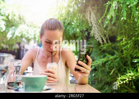 young urban woman sitting in street cafe drinking ice coffee using straw and holding mobile phone in her hands. Stock Photo