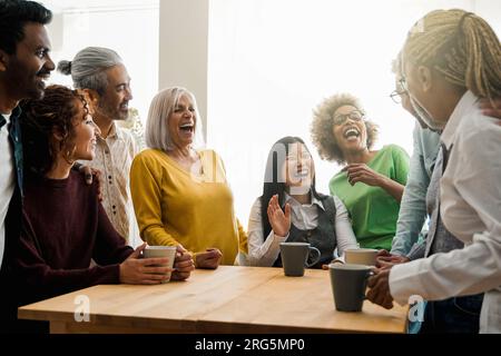 Happy multiethnic people drinking tea during lunch break at work - Group of multi-generational friends having fun laughing together - Soft focus on As Stock Photo