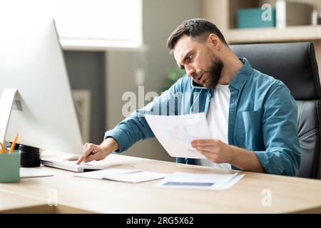 Multitasking. Young Businessman Working On Computer, Talking On Cellphone And Checking Documents Stock Photo