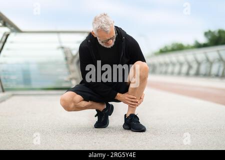 Senior man jogging by bridge, suffering from shin splint Stock Photo