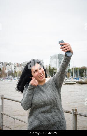 young latin woman on the pier over the river, wearing gray clothes, smiling happy taking a selfie with her phone with the sky in the background and co Stock Photo
