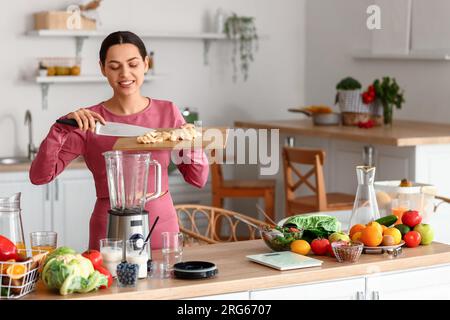 Sporty young woman putting cut banana into blender in kitchen Stock Photo