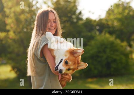 a young girl holds a cheerful and funny Welsh Corgi in her arms in a park in sunny weather, the concept of happy dogs Stock Photo