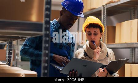 Multiethnic team of workers analyzing list on laptop and tablet, working with industrial cardboard boxes in storage room. People checking merchandise stock for delivery service. Handheld shot. Stock Photo