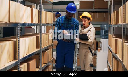 Multiethnic team of employees working with goods and checking stock inventory on laptop, cargo in storage room. Depot workers analyzing industrial goods to plan distribution and shipment. Stock Photo