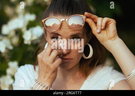 Summer time. sad modern 40 years old woman in white shirt with handkerchief and eyeglasses has an allergy attack near flowering tree. Stock Photo