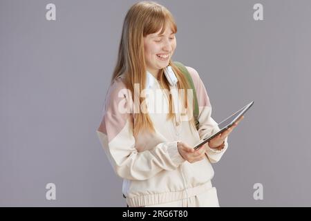 happy trendy teenager girl in beige tracksuit with headphones using tablet PC isolated on grey. Stock Photo