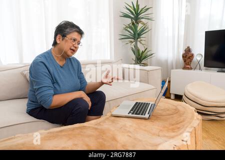 Elderly lady actively engaged with technology at home. She is surrounded by a cozy living room ambiance. Stock Photo