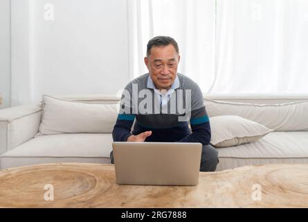 Ole man relaxing comfortably on a sofa, immersed in operating a laptop. Evident focus and concentration in a cozy domestic backdrop. Stock Photo