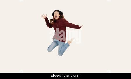 An overjoyed and pretty Asian woman in a red sweater is jumping with joy over an isolated white studio background. happy, cheerful, joyful, fun Stock Photo