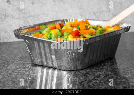 Mixed vegetables, with peas carrot sweetcorn broccoli and pepper, in a takeaway foil tray carton container. On a black granite background Stock Photo