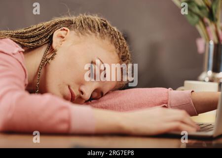 Young woman with box-braids falls asleep on laptop keyboard, hand on cellphone. Exhausted from work or study Stock Photo