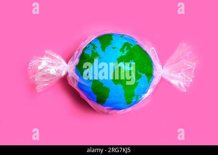 Miniature globe wrapped in plastic packaging like a candy, isolated on pink background. Planet plastic pollution related concept. Stock Photo