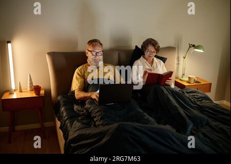 Happy retired family couple in bed using laptop and reading book Stock Photo