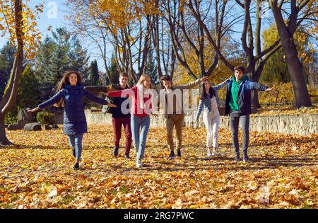 Happy smiling friends enjoy walking through the autumn park, running with their arms outstretched to sides on sunny warm day. Stock Photo
