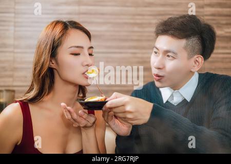 Young asian man feeding happy girlfriend with tasty food in restaurant Stock Photo
