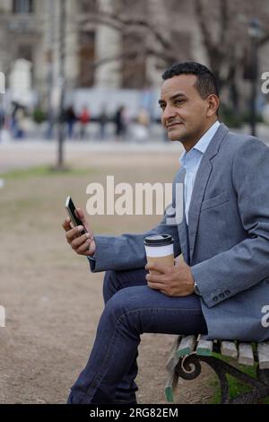 Portrait of latino man in jacket with a disposable cup of coffee sitting on a bench in a public park. Stock Photo