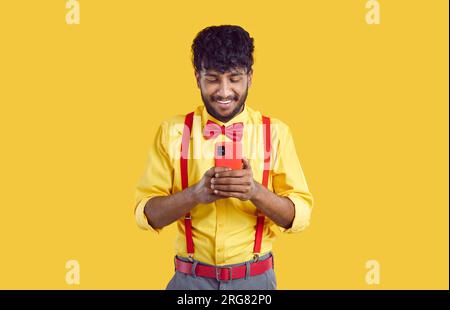 Happy young Indian man standing on yellow background, looking at cellphone and smiling Stock Photo