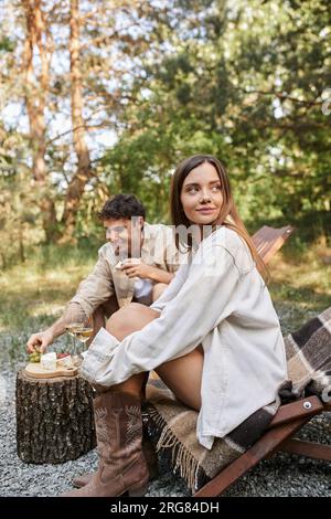 Brunette woman sitting on deck chair near boyfriend with wine and food during picnic outdoors Stock Photo