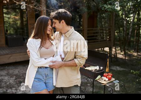 Joyful romantic couple hugging while standing near bbq and vacation house during picnic outdoors Stock Photo