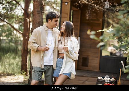 Smiling couple with wine hugging near grill and blurred vacation house during picnic at background Stock Photo