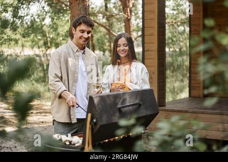 Smiling couple with wine cooking on grill while resting near vacation house at background Stock Photo