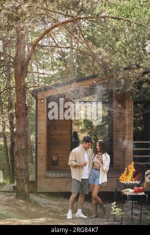 Smiling and stylish couple with wine hugging near bbq during picnic near vacation house outdoors Stock Photo