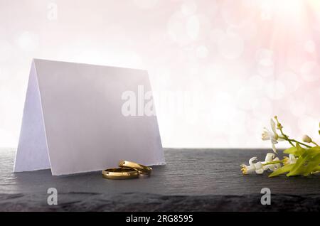 Two engagement rings on slate table with blank paper sign behind and flower bouquet with light pink background and bokeh effect. Front view. Stock Photo