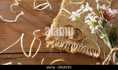 Detail of two gold rings on sackcloth on a rustic wooden table with straw and a bouquet of flowers. Top view. Stock Photo