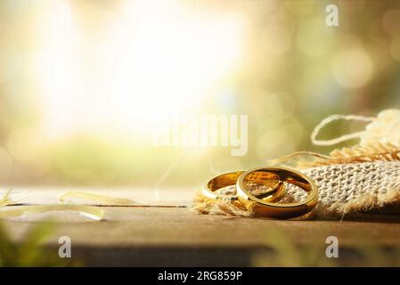 Detail of two gold rings on sackcloth on a rustic wooden table and green nature background with bokeh effect. Front view. Stock Photo