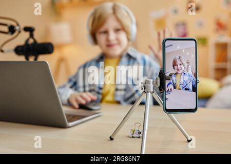 Focus on smartphone screen with cute schoolboy clicking mouse while sitting by desks in front of gadgets and recording new video Stock Photo