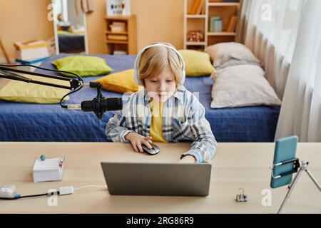 Blond schoolboy in casualwear and headphones playing sport video game or making livestream while sitting by desk in front of mobile gadgets Stock Photo
