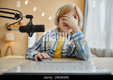 Upset or scared little boy in headphones covering his face with hand while sitting in front of computer and microphone and clicking mouse Stock Photo