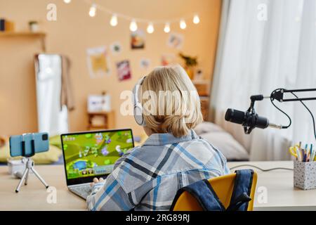 Rear view of blond schoolboy typing on laptop keyboard while sitting by desk in front of camera in home environment and playing video game Stock Photo