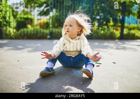 Adorable toddler girl with dirty hands after drawing with colorful chalks on asphalt. Outdoor activity and creative games for small kids Stock Photo