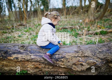 Adorable preschooler girl sitting on log and having fun in spring forest or park. Child enjoying nature on a spring day. Outdoor activities for kids Stock Photo