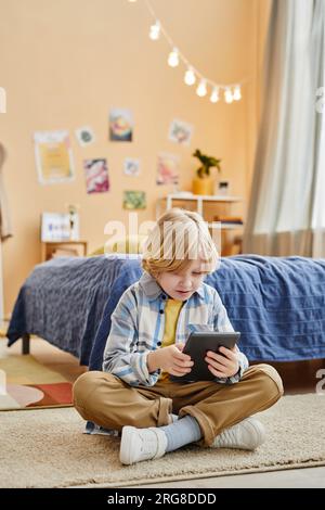 Cute blond boy looking at screen of table while sitting on the floor by comfortable bed and watching online movie, cartoon or video Stock Photo