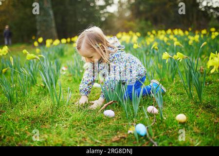 Three year old girl playing egg hunt on Easter. Child sitting on the grass with many narcissi and gathering colorful eggs in basket. Little kid celebr Stock Photo