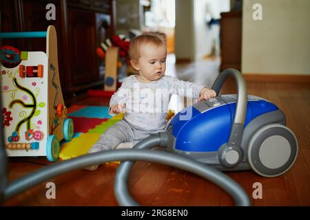Little baby girl playing with vacuum cleaner at home. Toddler helping parents with chores. Stock Photo