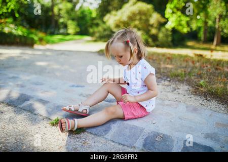 Cute little toddler girl sitting on the ground after falling at summer park. Child getting hurt while walking in park. Kid looking at her boo-boo Stock Photo