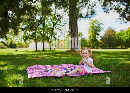 Adorable toddler girl having picnic in countryside on a summer day. Child enjoying healthy snack in park. Outdoor summer activity for small kids Stock Photo
