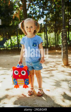 Adorable toddler girl with funny backpack ready to go to daycare, kindergarten or school. First day of school concept Stock Photo