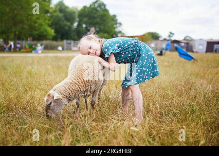 Adorable preschooler girl playing with sheep at farm. Child familiarizing herself with animals. Farming and gardening for small children. Outdoor summ Stock Photo