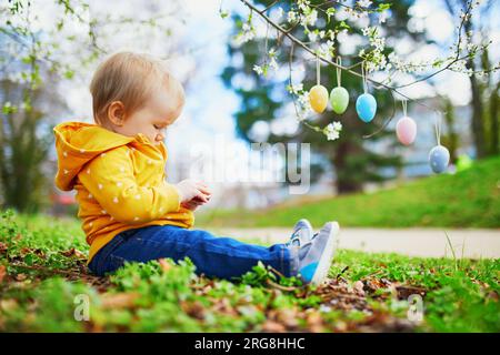 Cute little one year old girl playing egg hunt on Easter. Toddler sitting on the grass under apple tree in full bloom decorated with colorful eggs. Li Stock Photo