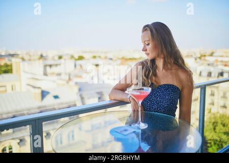 Beautiful young woman drinking alcoholic cocktail in a rooftop restaurant with view to the Eiffel tower in Paris, France Stock Photo
