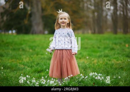 Cute preschooler girl in princess crown standing in the grass with many snowdrop flowers in park or forest on a spring day. Little kid exploring natur Stock Photo