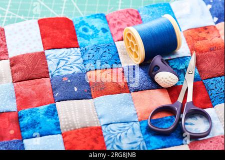 Scissors, thread and thimble lying on blue and red square pieces of fabric sewn together Stock Photo