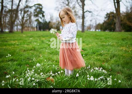 Cute preschooler girl in pink tutu skirt standing in the grass with many snowdrop flowers in park or forest on a spring day. Little kid exploring natu Stock Photo